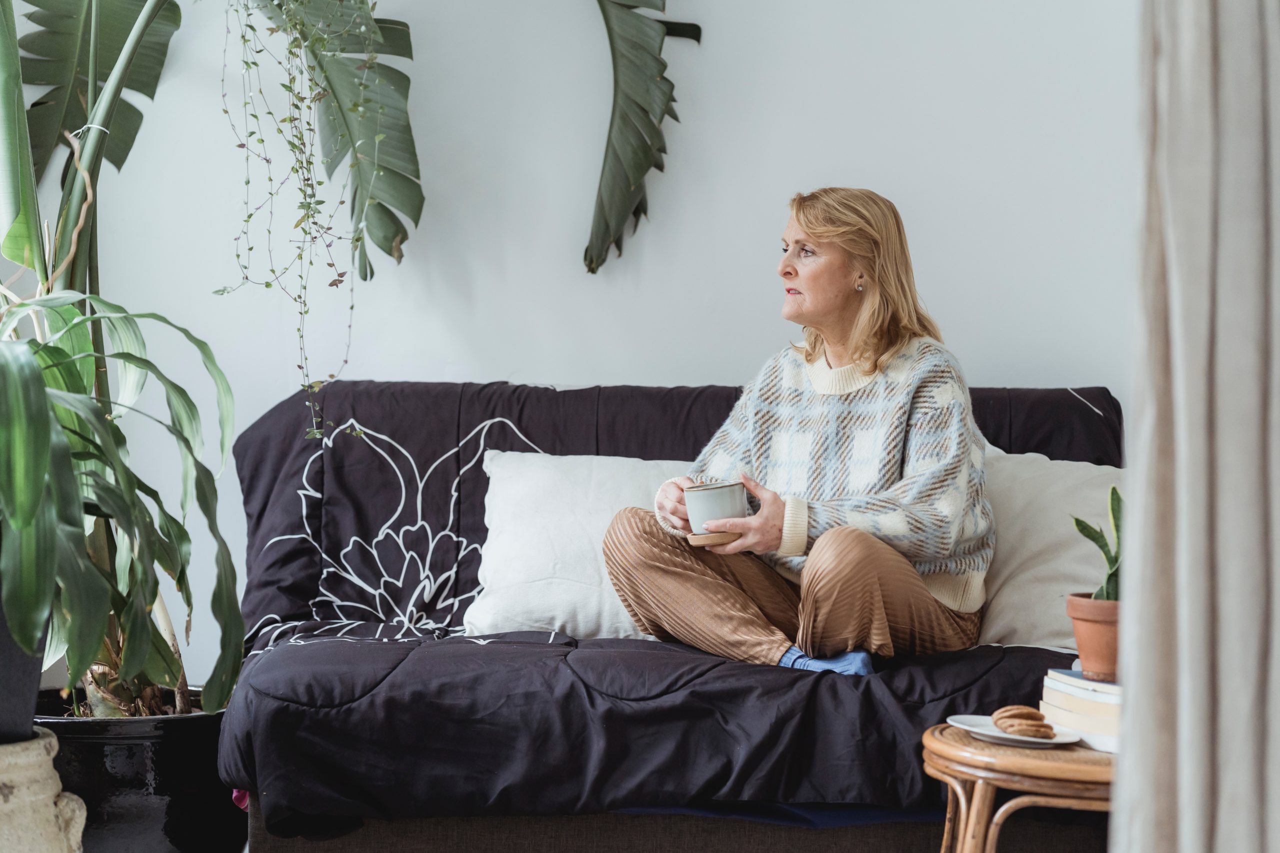Thoughtful mature woman sitting on couch with crossed legs and drinking hot beverage